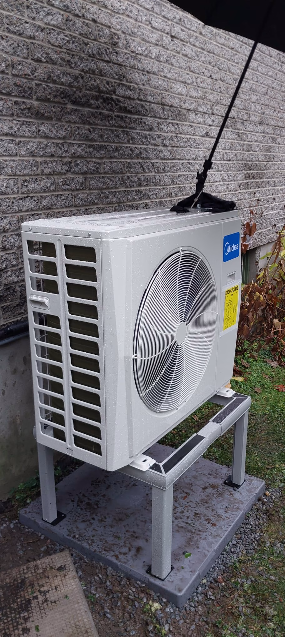 Midea outdoor heat pump on a raised stand beside a masonry wall.